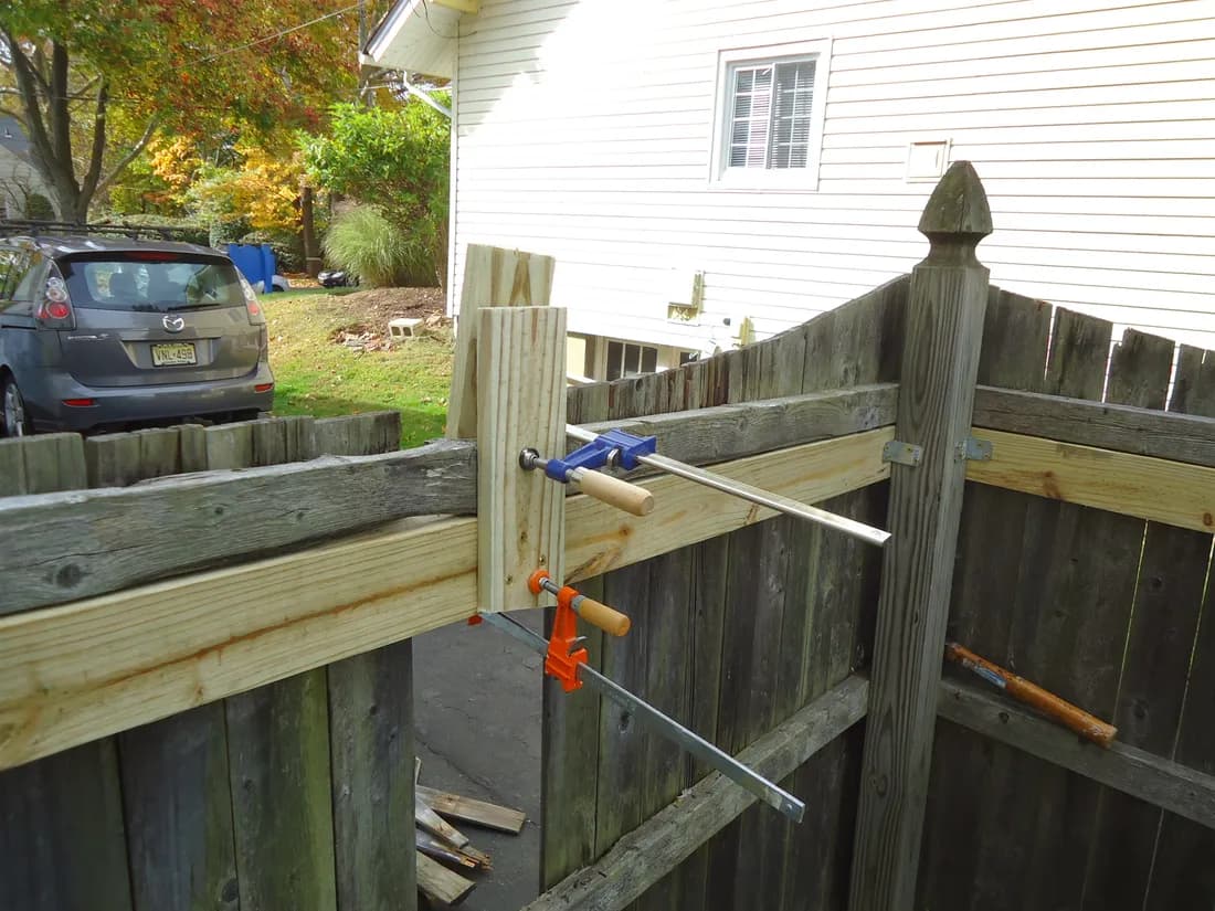 Hands clamping new lumber onto an existing wood fence during a repair